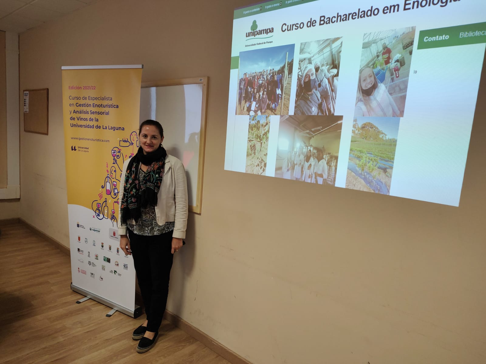 Professora Ângela ministra curso de Enoturismo sobre vitivinicultura brasileira na Universidade de La Laguna na Espanha Foto colorida da Professora Ângela Marcon à esquerda, ela está ao lado de cartaz do curso sobre enoturismo ministrado na Espanha