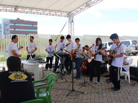 A orquestra brindou o espaço da reflexão com melodias no primeiro plano, um homem está sentado, de costas para a câmera, e olha para os integrantes da orquestra, com diversos instrumentos musicais. Eles estão em pé, usam camisetas claras e tocam música.