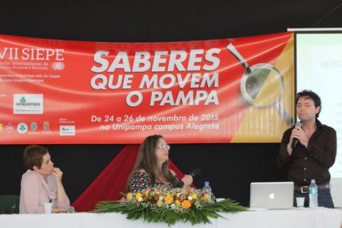 Mesa-redonda tratou das experiências e do valor do aprendizado de outros idiomas duas mulheres estão sentadas à frente da mesa de honra, e olham para um homem que fala ao microfone. Ao fundo, um banner com a marca e o nome do evento