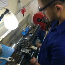 Infraestrutura permite ligar saberes científicos e problemas práticos de engenharia mecânica A foto mostra uma bancada com equipamento de torno e lâmpada, na qual um aluno prepara uma peça para uso em um projeto