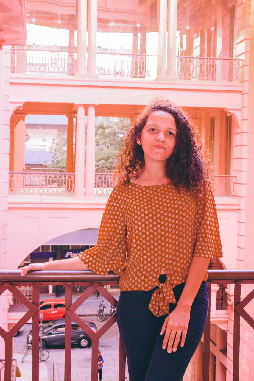 Menção coroa trabalho feito pela egressa da Unipampa em evento nacional - Divulgação Foto colorida da egressa Anna Carolina Furlanetto no canto direito da imagem, ela tem pele branca, cabelo escuro cacheado