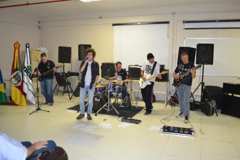 Unipampa Band Rock animou primeira noite do PampaPET Três bandeiras, a do Brasil, do Rio Grande do Sul e da Unipampa aparecem no fundo, à esquerda. Uma banda formada por cinco rapazes e que conta com bateria, baixo, guitarra e vocal se apresenta dentro de um auditório, em frente a um quadro branco usado em aulas